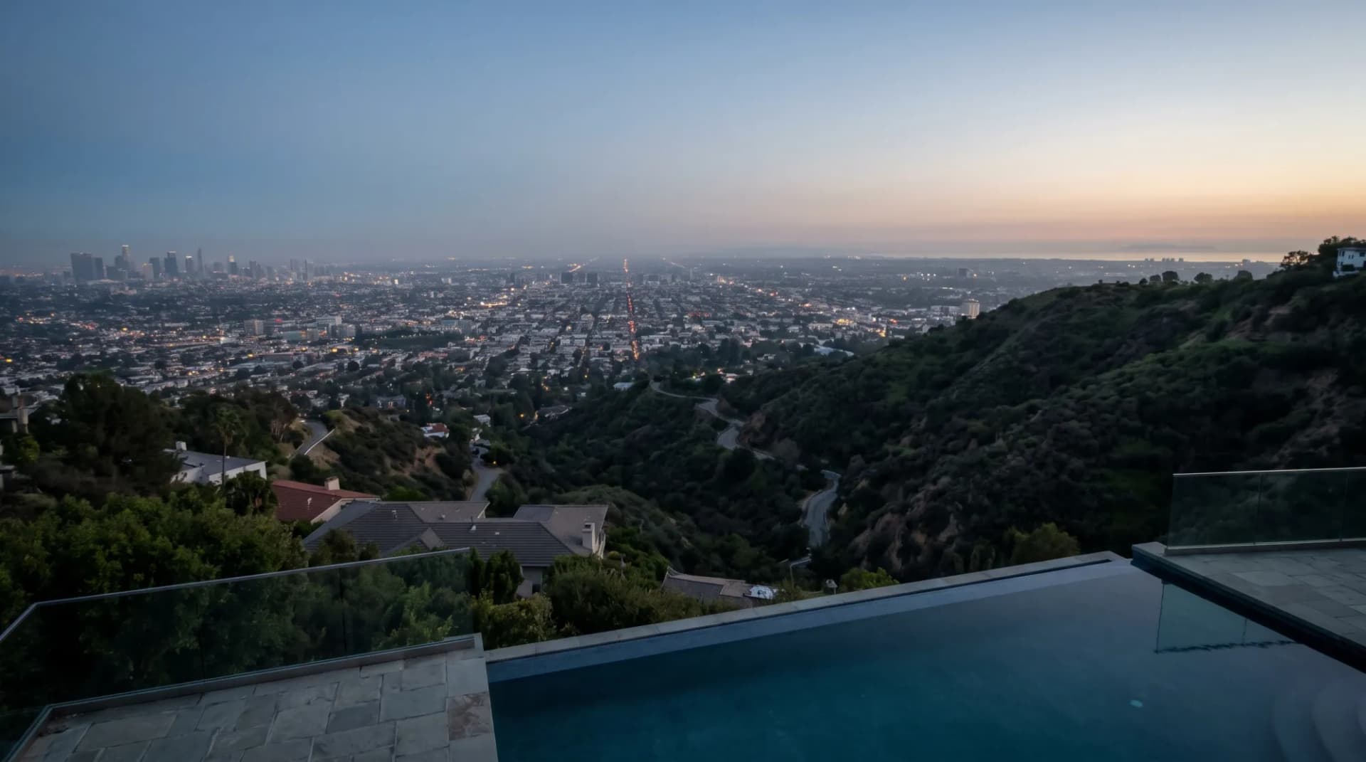 A hilltop infinity pool terrace at dusk overlooking the lights of Los Angeles, with downtown on one side and the ocean on the other.