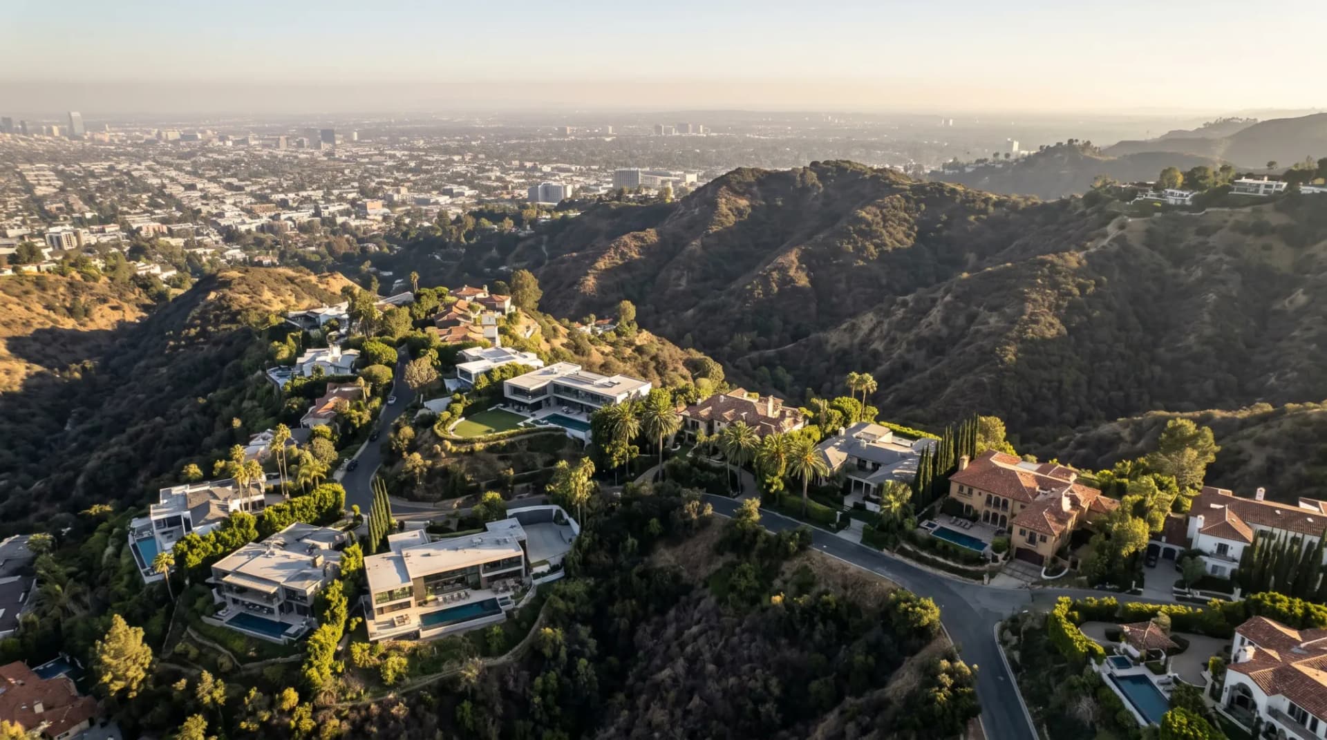 Aerial view of hillside luxury estates with pools above the hazy city grid of west Los Angeles in late-afternoon light.